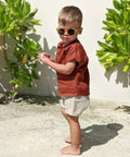 Child wearing sunglasses and a red shirt standing outdoors with green plants in the background