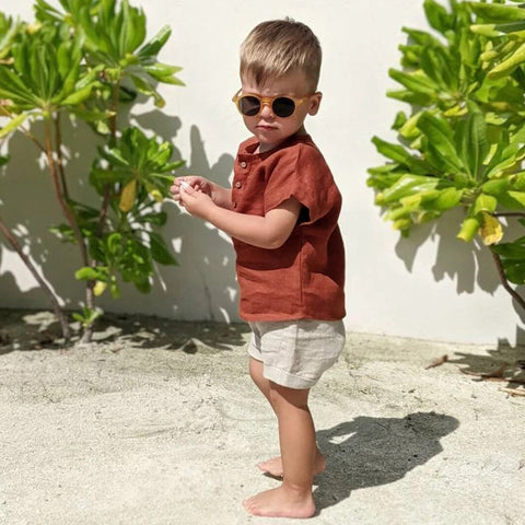 Child wearing sunglasses and a red shirt standing outdoors with green plants in the background