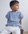 Child wearing a blue and white striped shirt sitting on a woven stool against a plain background