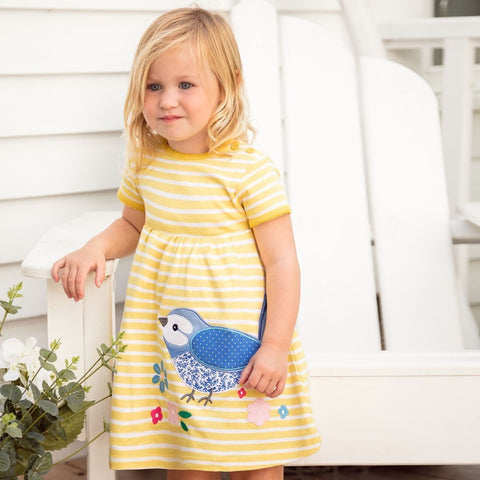 Young girl wearing a yellow striped dress with a blue bird design, standing on a white surface.