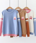 Three sweaters in blue, brown, and white with pink and blue accents on wooden hangers against a white wall.
