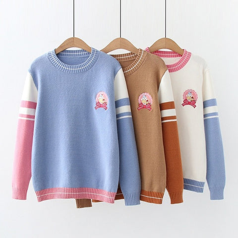Three sweaters in blue, brown, and white with pink and blue accents on wooden hangers against a white wall.