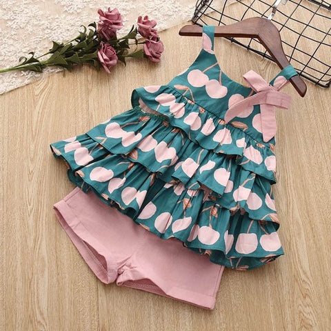 Children's dress with cherry pattern and pink shorts on a wooden surface.
