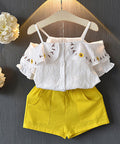 White embroidered top and yellow shorts set on a hanger against a dark background with flowers.