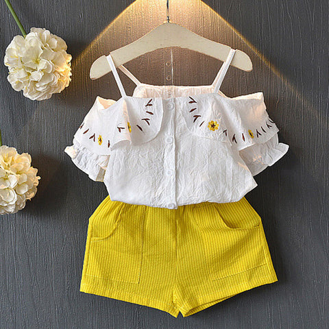 White embroidered top and yellow shorts set on a hanger against a dark background with flowers.