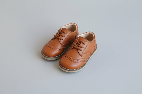 Pair of brown children's shoes on a white surface with a hand holding one shoe.