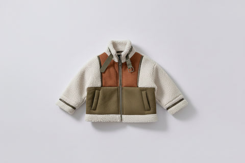 Children's multicolored jacket with a hanger on a white surface, with a red bag and plate of food in the background.