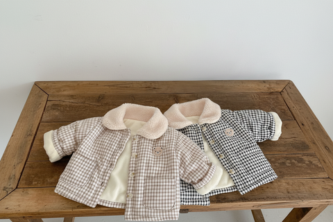 Two children's jackets with checkered pattern and pink fur collar on a wooden surface with an open book.