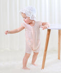 Child wearing a light pink outfit and white bonnet standing on a white floor with a white curtain background.