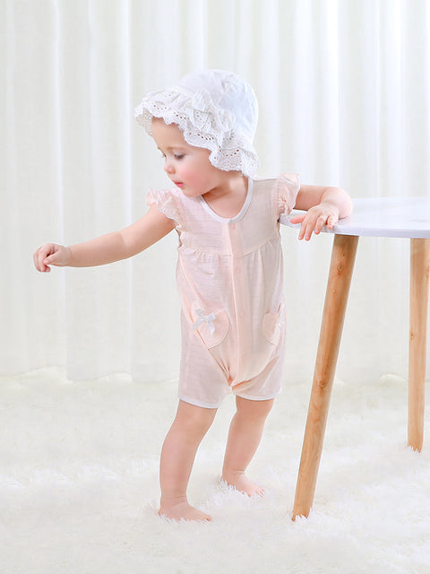 Child wearing a light pink outfit and white bonnet standing on a white floor with a white curtain background.