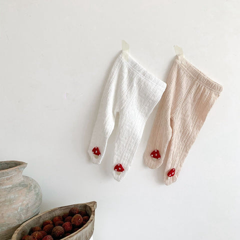 Two pairs of baby leggings with red accents on a white surface, next to a ceramic pot and bowl with berries.