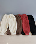 Four pairs of children's corduroy pants in beige, brown, red, and black on a light wooden floor.