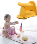 Baby sitting on a pink bath seat with a yellow bathtub support, playing with toys on a white mat.