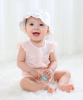Baby sitting on a white surface wearing a white bonnet and pink outfit.