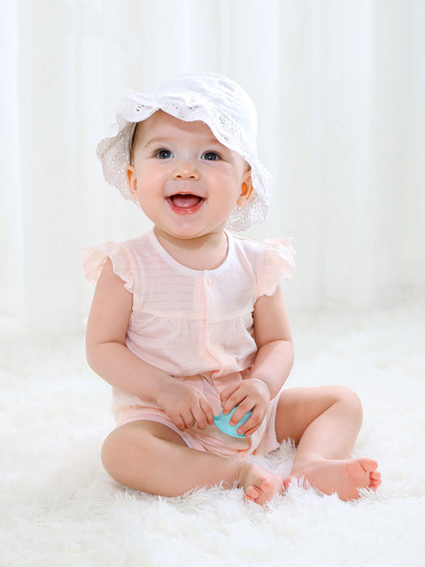 Baby sitting on a white surface wearing a white bonnet and pink outfit.