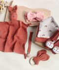 Collection of baby items including a pink rabbit plush, bow headbands, a muslin blanket, and toys on a light background.