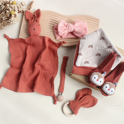Collection of baby items including a pink rabbit plush, bow headbands, a muslin blanket, and toys on a light background.