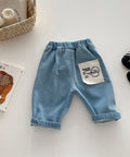 Blue denim baby pants with a pocket on a light surface, surrounded by a book, basket, and shoes.