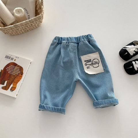Blue denim baby pants with a pocket on a light surface, surrounded by a book, basket, and shoes.
