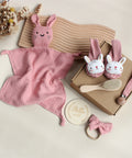 Baby care products including a pink knitted blanket with bunny ears, baby booties, a hairbrush, and a 'Hello world' coaster on a neutral background.
