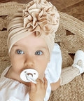 Baby wearing a beige turban with a flower and holding a white pacifier, lying on a woven surface.