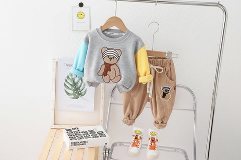 Children's clothing set with gray shirt, yellow sleeves, brown pants, and shoes on hangers against a white wall.