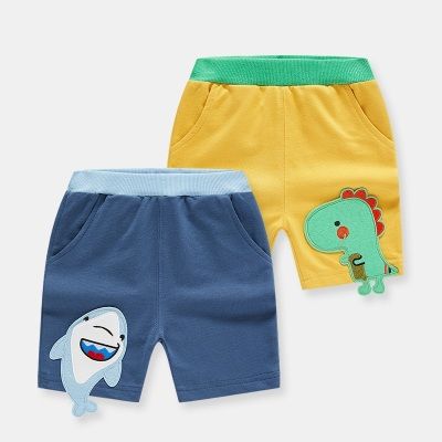 Two pairs of children's shorts, one blue with a shark design and one yellow with a dinosaur design, on a light gray background.