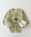 Children's green sweatshirt with teddy bear graphic and 'California' text, paired with a knit hat and white shoes on a light background.