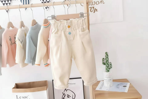 Beige pants with colorful buttons on a hanger against a white wall.